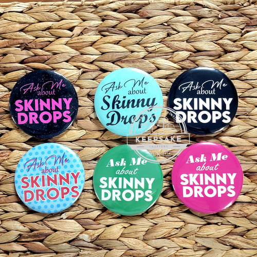 A collection of colorful pins with the text 'Ask Me About Skinny Drops' displayed on them, placed on a woven straw surface.