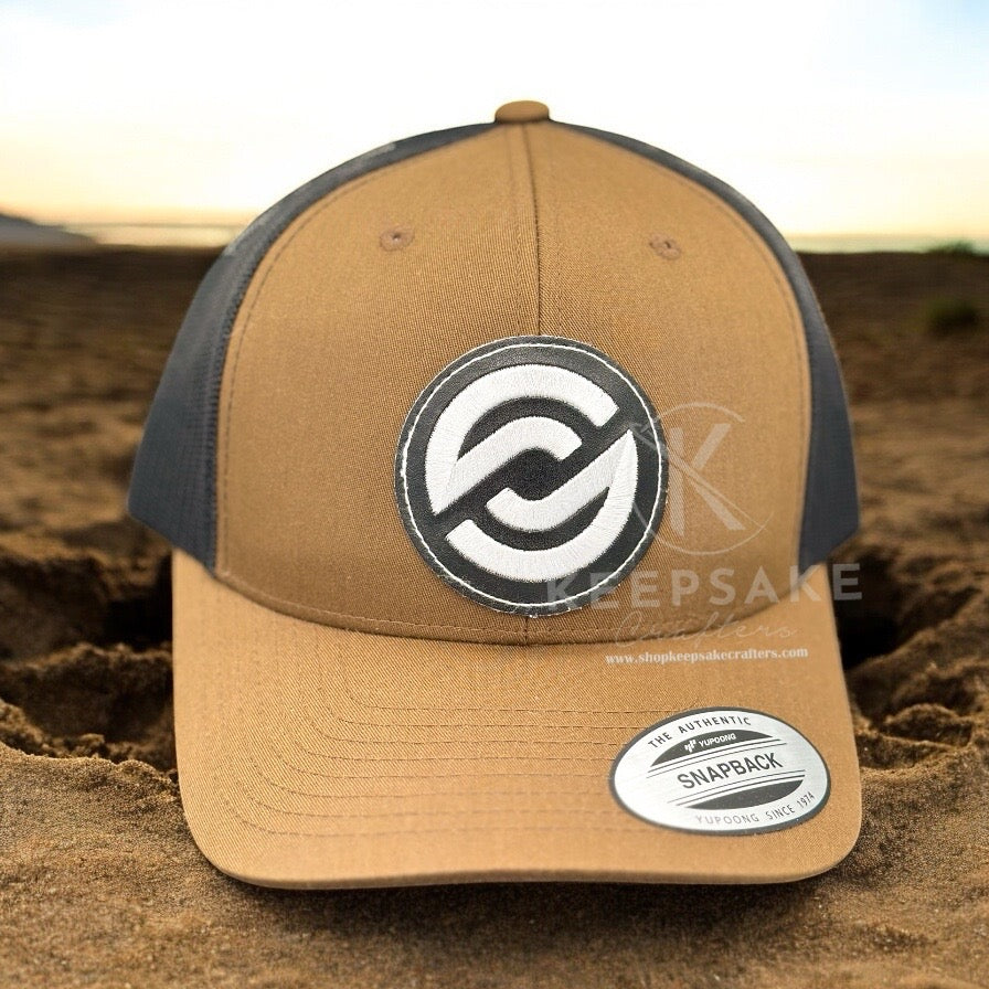 A brown and black trucker cap with a white embroidered design on the front, displayed on a sandy surface.