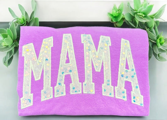 Neon violet T-shirt with the word 'MAMA' customized in white sparkly sequins, displayed on a flat surface with green plants in the background.